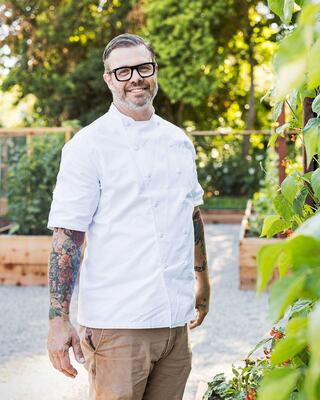 Smiling Chef Standing in Garden with Short-Sleeve Chef Coat, Glasses, and Arm Tattoos