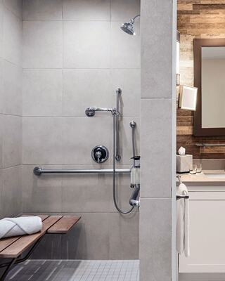 An Accessible Hotel Room with a Walk In Shower with a Bench and Handrail, and a Bathroom Vanity Next to it