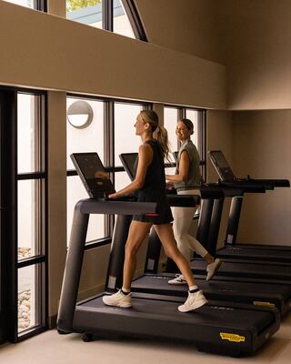 Two women are jogging on treadmills in a bright, modern exercise space, enjoying their workout with large windows and natural light.