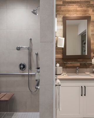An Accessible Hotel Room with a Walk In Shower with a Bench and Handrail, and a Bathroom Vanity Next to it
